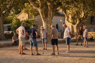 Group of pétanque players talking and watching over a cochonnet on a shaded outdoor boulodrome lined with plane trees, with sunlight and Provençal village houses in the background.