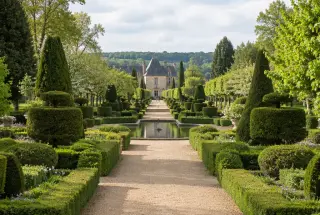 Allée centrale bordée de topiaires et de haies aux Jardins d’Eyrignac, avec un bassin et une perspective vers un bâtiment en pierre.